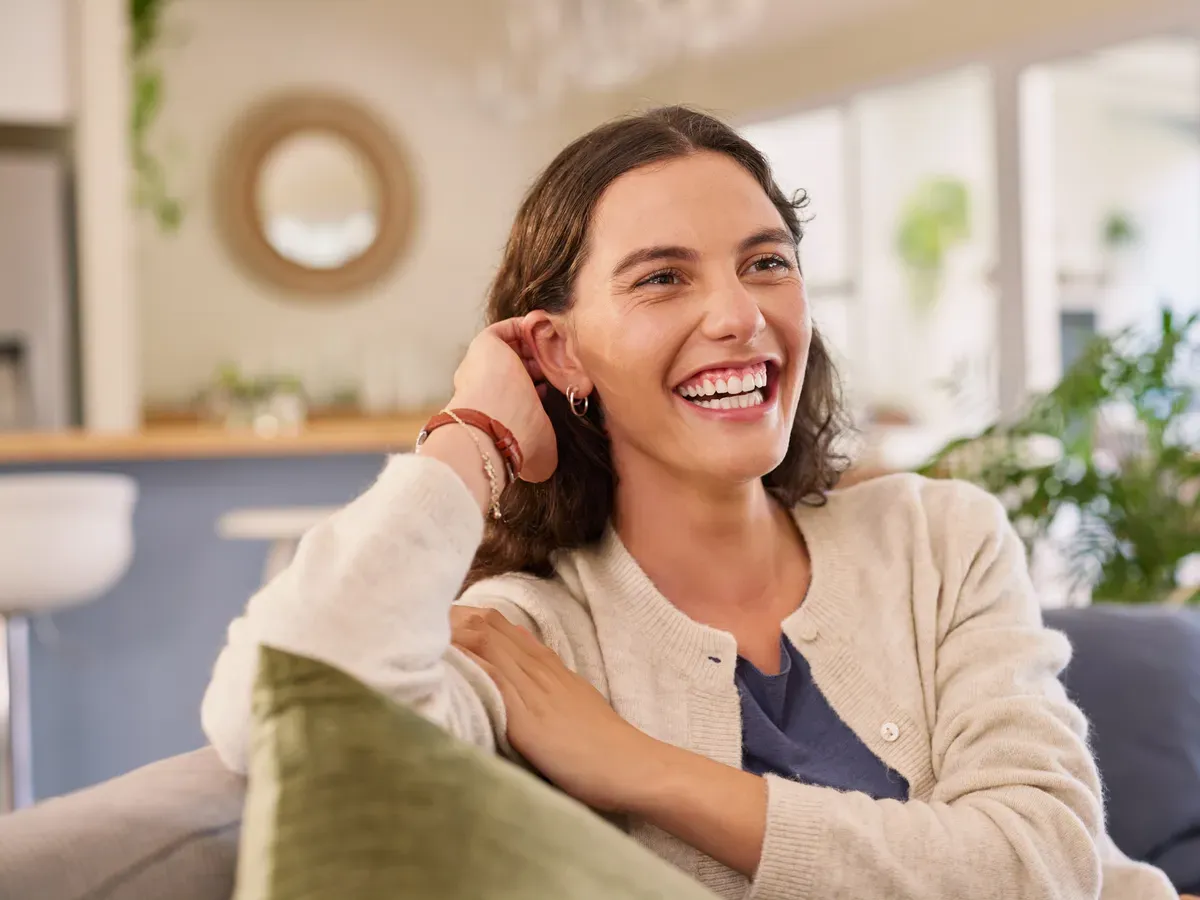 A woman sits on the couch at home, laughing during a video appointment