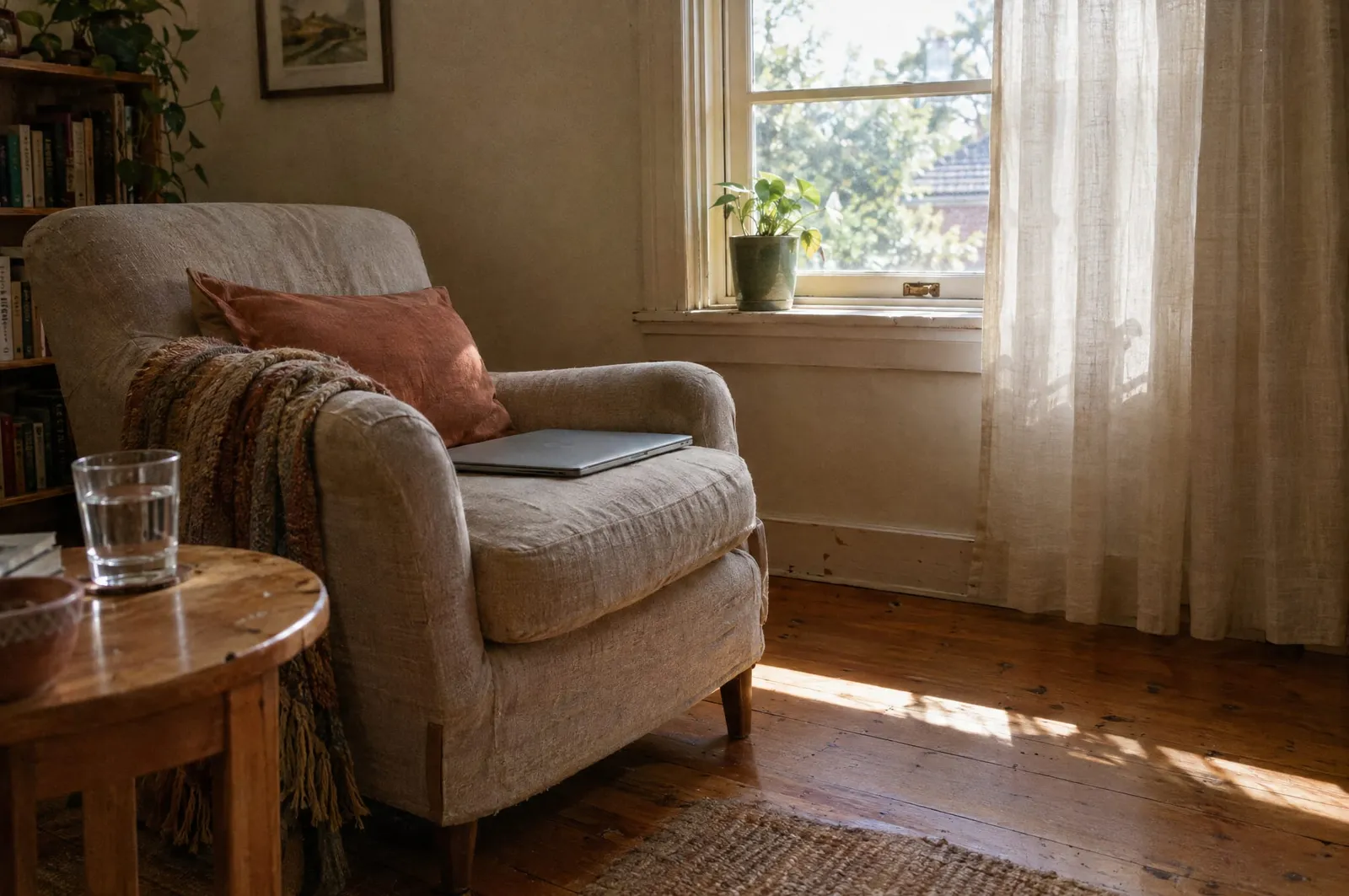 An empty linen armchair beside a sunlit window in a home living room, a closed laptop on the seat and a glass of water on the side table