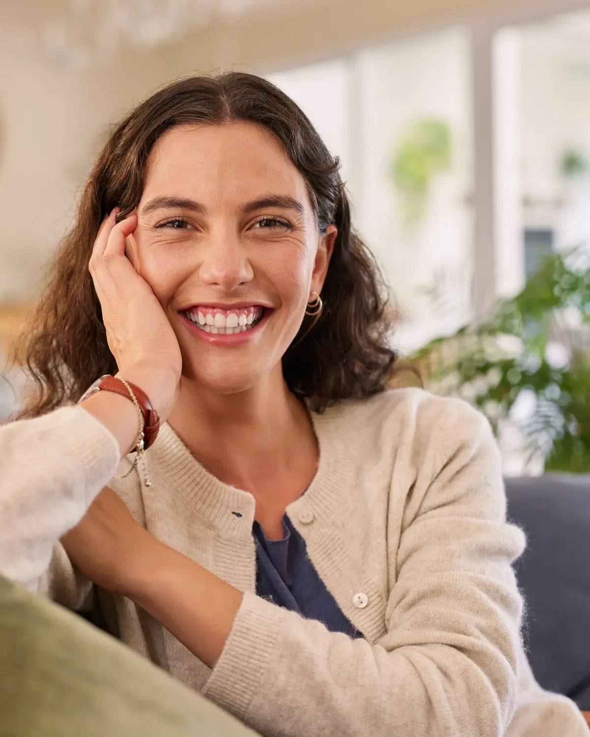 A woman sits on the couch at home, resting her chin on her hand during a video appointment